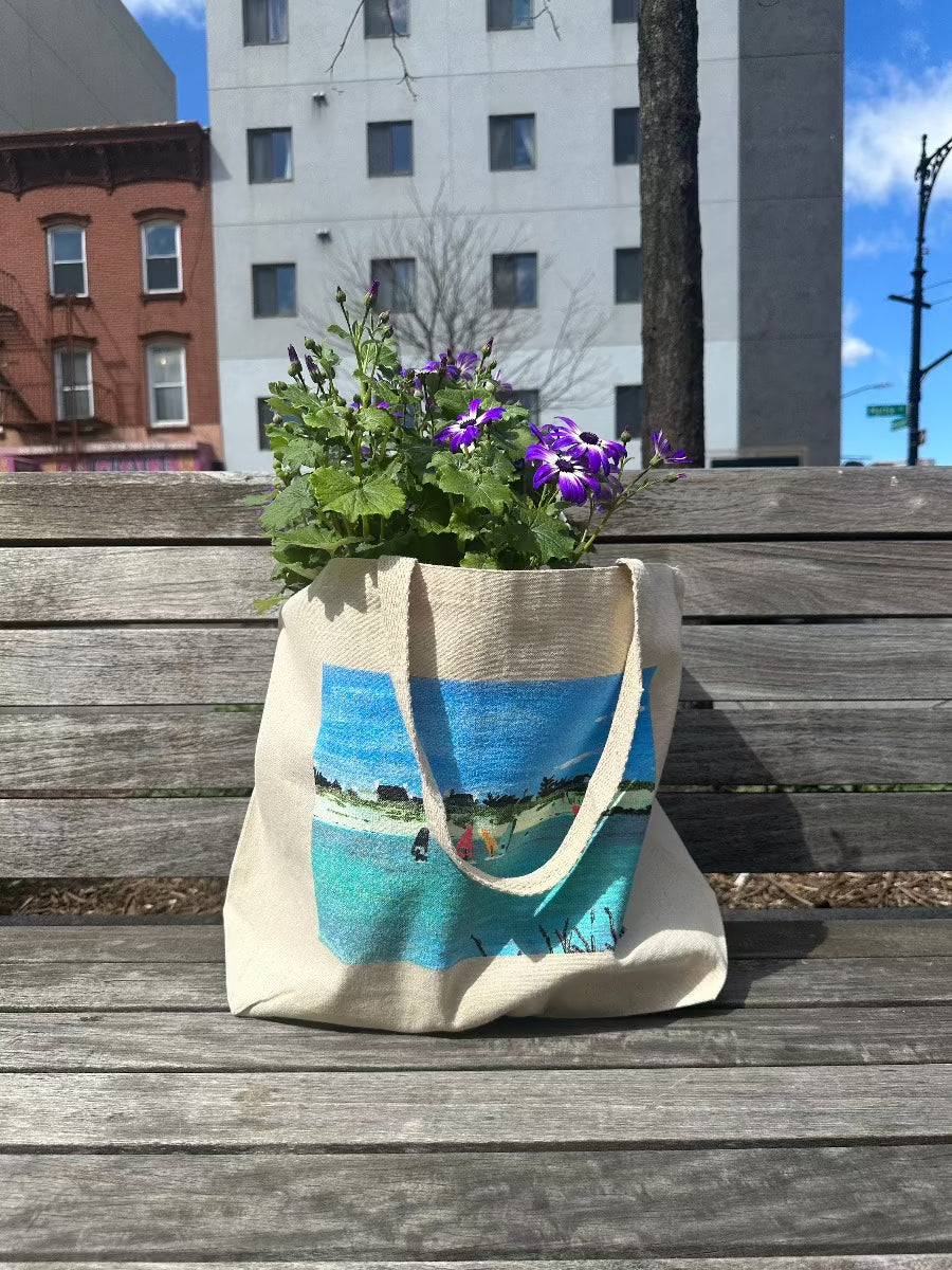 Tote bag with boats on water scene. Bag has purple flowers in it, sitting on park bench in city