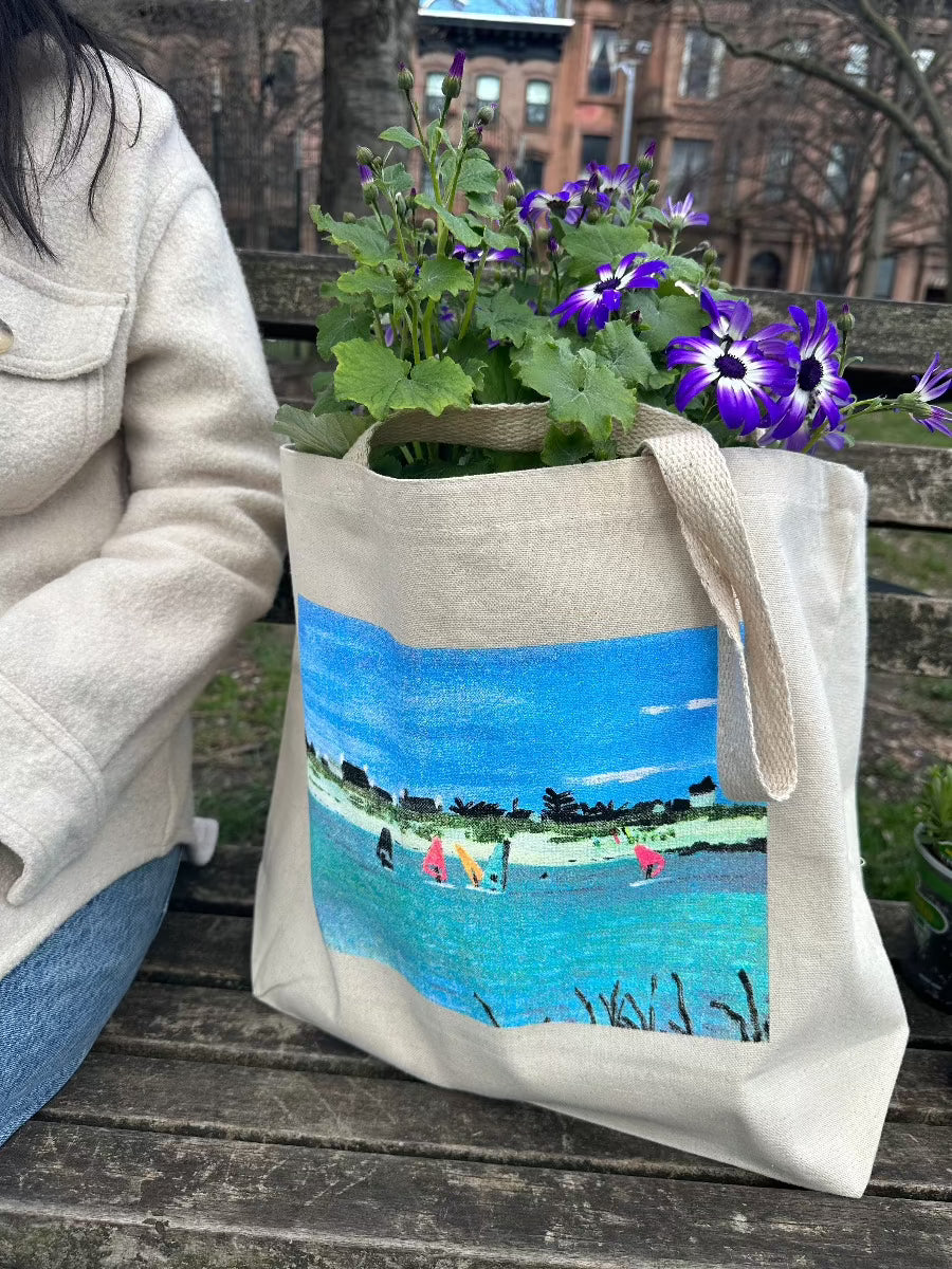 Tote bag with boats on water scene. Bag has purple flowers in it, sitting on park bench in city