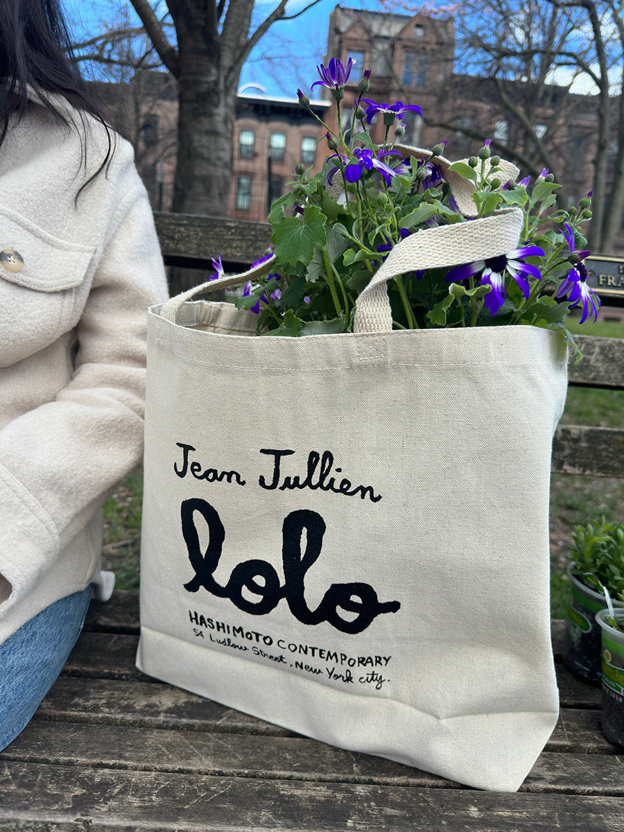 Tote bag with text "Jean Jullien Lolo Hashimoto Contemporary 54 Ludlow Street New York City'. Bag has purple flowers in it, sitting on park bench in city