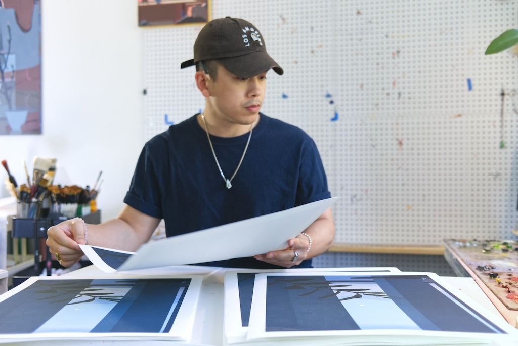 Person holding a large sheet of paper in an art studio with framed artwork on the wall.