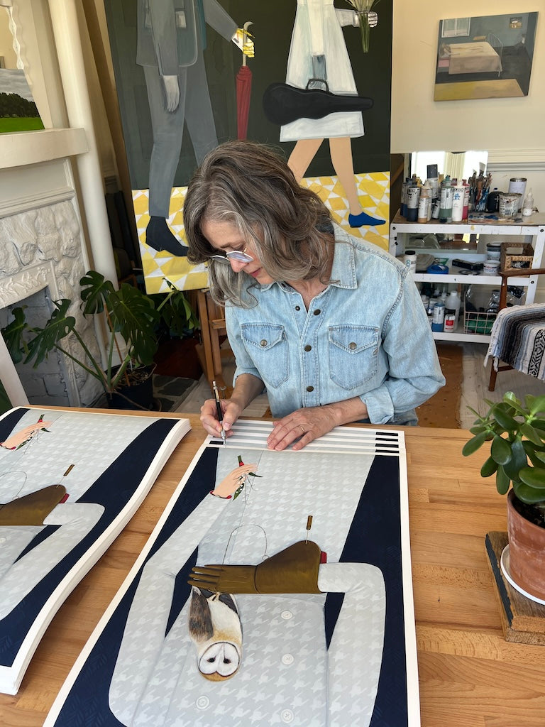 woman in blue shirt wearing glasses signing print of a woman's torso in a blue dress holding a owl in her brown gloved hand