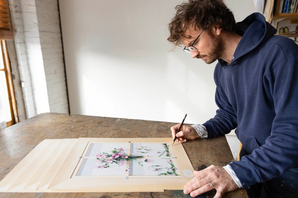 Stephen Morrison (white man with glasses in blue hoodie) signing print sof trompe-l'oiel back of canvas (wooden stretcher bars) with flowers at center and falling. Flowers have small dog face at center