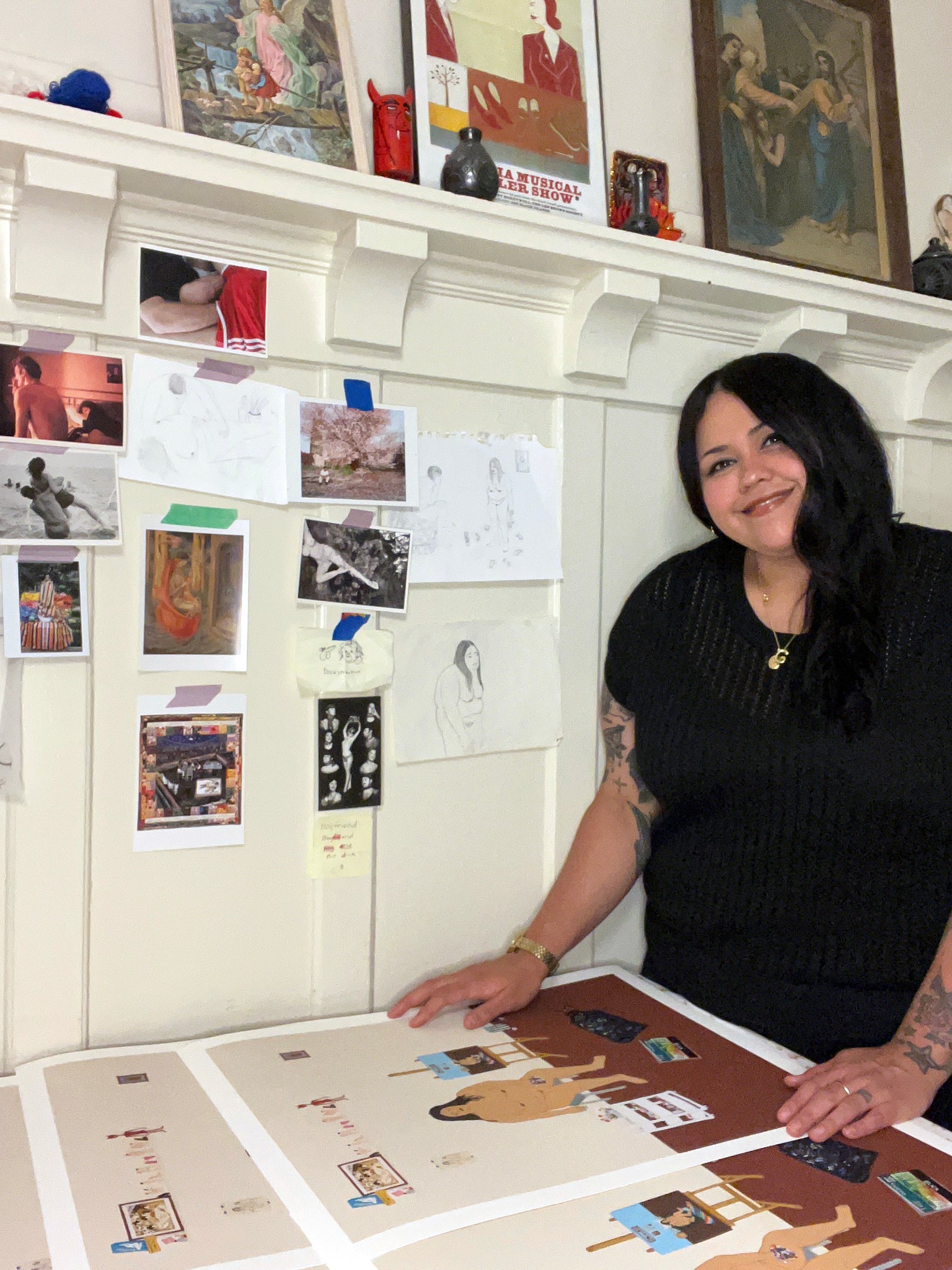 artist Gina M. Contreras smiling at camera (woman with dark hair in black tshirt) standing over prints of a large nude woman in a beige room, seated in front of an easel with a painting in progress. The room is relatively bare besides a shelf with many erotic objects and tchotchkes, and miscellaneous art supplies. Artist references hanging on wall behind her