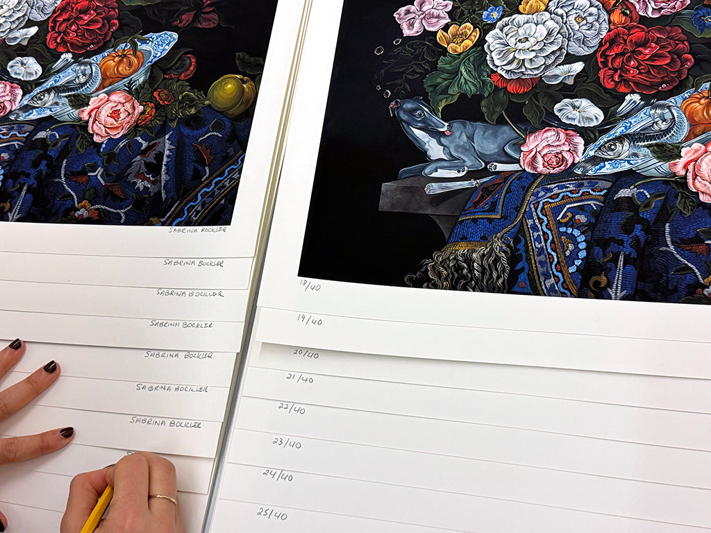 Sabrina Bockler signing print of a still life featuring a bouquet of flowers, a grey dog, and an ornately patterned blue cloth against a black background by Sabrina Bockler