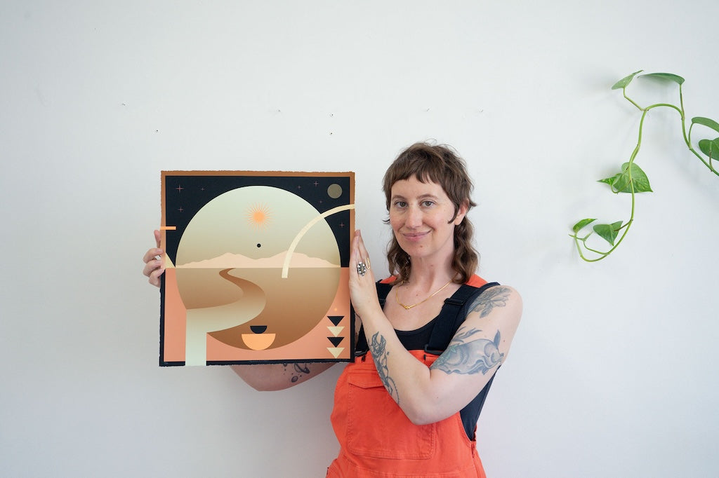 Portrait of white woman holding up summer solstice inspired print in studio; green plant hanging on right